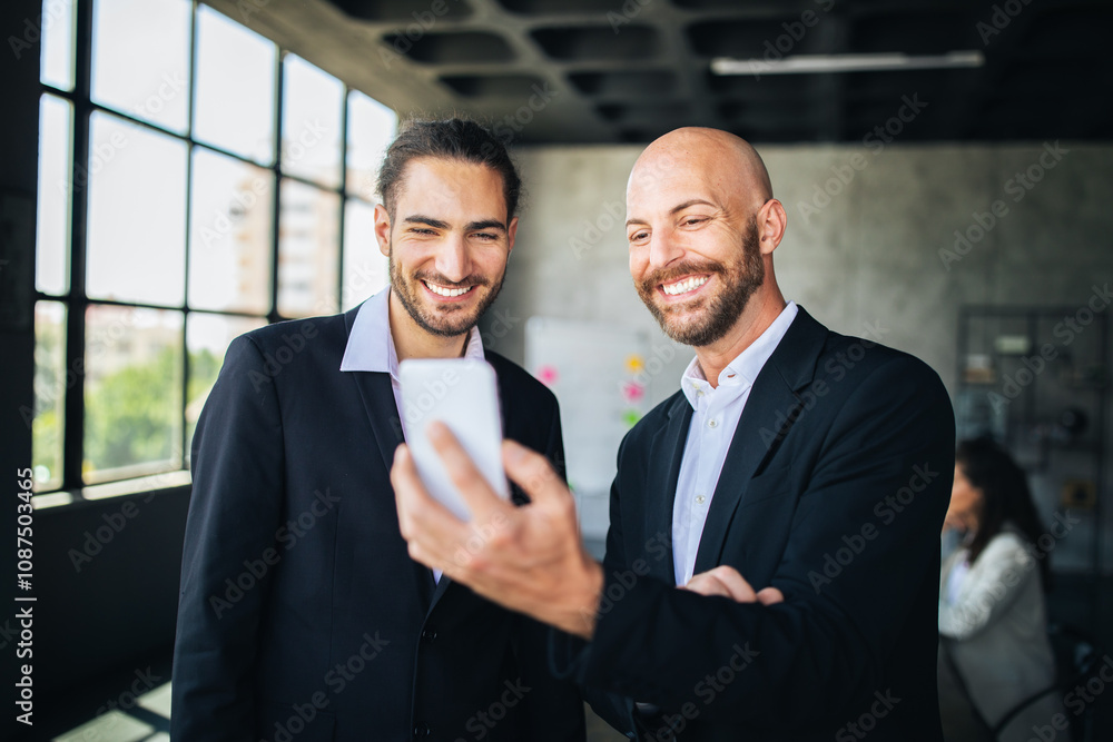 Leinwandbilder two men in formal attire are smiling at a smartphone screen in a modern office. natural light enters