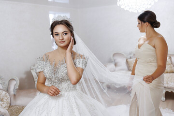 A bride is getting ready for her wedding, and her bridesmaid is helping her. The bride is wearing a...
