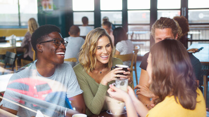 Multi-Cultural Group Of Friends Sitting At Table By Window In Busy Coffee Shop Talking