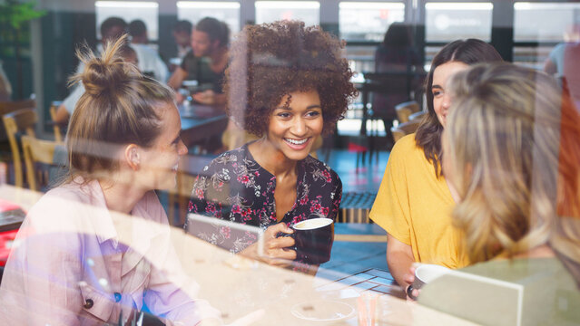 Multi-Cultural Group Of Female Friends Sitting At Table By Window In Busy Coffee Shop Talking