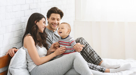 Cheerful millennial man and woman playing with baby son, sitting together on bed, free space