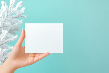Obraz premium A hand holding a blank white card in front of frosted pine branches, set against a light blue background in a wintery theme.