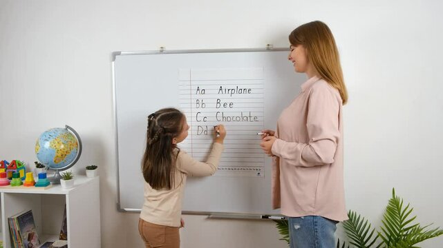 A young girl is learning to write on a whiteboard with the help of her teacher. They are both smiling and happy. The classroom is bright and cheerful, with a globe and toys on a table.