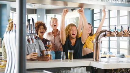 Group Of Young Male And Female Friends Celebrating Whilst Watching Game On Screen In Sports Bar