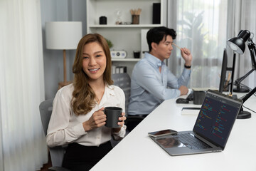 Profile smiling beautiful Asian businesswoman looking camera while holding cup to pose at modern office on working desk. Blurry background man colleague analyzing data market report on pc. Infobahn.