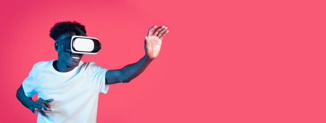 A young black man is fully immersed in a virtual reality experience, wearing a headset and smiling widely as he interacts with an unseen digital environment, surrounded by a vivid pink backdrop.
