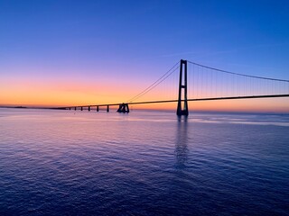 a stunning sunset view of the Great Belt Bridge (Storebæltsbroen) in Denmark. The sky is painted in hues of orange, pink, and blue. The bridge extends gracefully across the horizon. 
