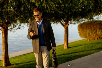 A stylish man enjoys a leisurely stroll along the waterfront, chatting on his phone amidst autumn foliage in the golden afternoon light