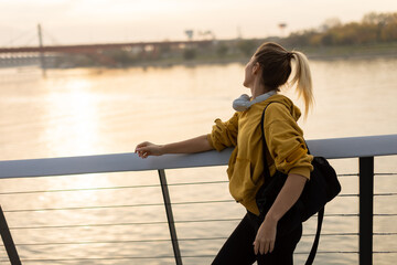 A joyful woman strolls along a modern riverside promenade at sunset, carrying a stylish bag and...