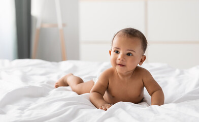 Cute Smiling African Baby Lying On Stomach Looking Away In Bedroom At Home. Naked Toddler Boy...