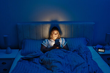 A young woman is using her smartphone while relaxing in bed during the night hours