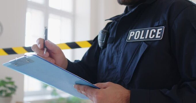 Cropped shot of male police officer collecting evidence at crime scene, interviewing witness and taking notes on clipboard, red blue lights flashing on his uniform. Police work, investigation concept