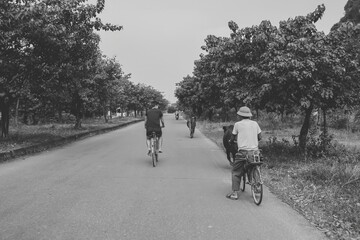A black-and-white photograph captures two men riding bicycles down a quiet rural road lined with trees.