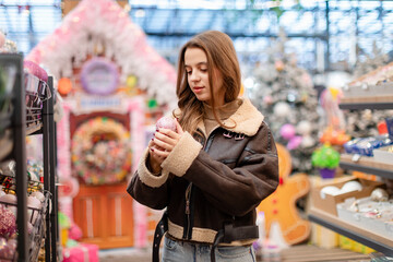 A young woman with long hair, wearing a short brown sheepskin coat, is choosing Christmas ornaments...