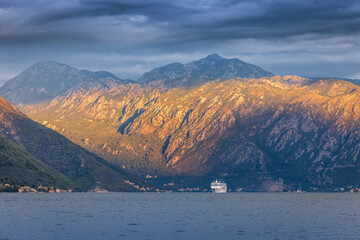 A serene view of the Bay of Kotor in Montenegro, featuring a white ship gliding across calm waters under a cloudy sky, with rugged, sunlit mountains in the background.