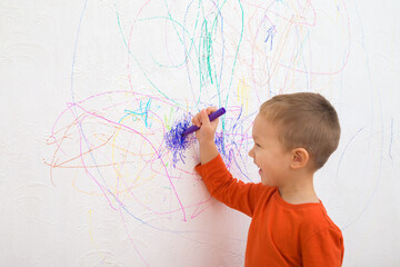 Happy smiling baby boy holding marker and drawing colorful scratches lines on white wallpaper background. Closeup. Draw of naughty little kid. Side view.
