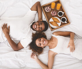 Honeymoon concept. Happy black young couple lying in bed in morning with breakfast, top view