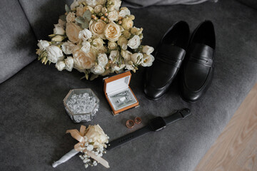 A man's wedding shoes and a bouquet of flowers are displayed on a couch. The flowers are white and the shoes are black