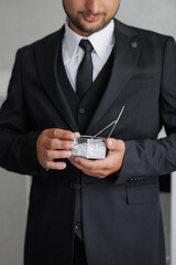 A man in a suit holding a small box with a silver chain. The man is wearing a black tie and a black vest