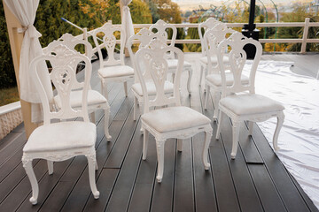 A row of white chairs are arranged on a wooden deck. The chairs are all facing the same direction, and they are all empty. The scene gives off a sense of calm and serenity
