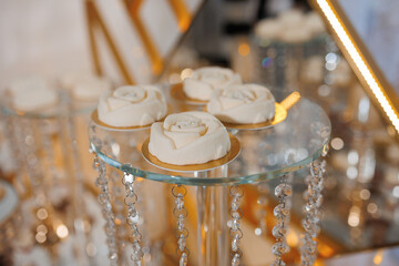 A glass display case with four small white cakes on top of it. The cakes are decorated with a rose design and are placed on a silver stand. The display case is surrounded by a clear glass base