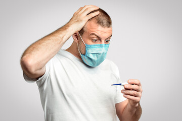 Fever Concept. Shocked Sick Man In Medical Mask Looking At Thermometer Having Coronavirus Symptom Standing Over Gray Background. Panorama