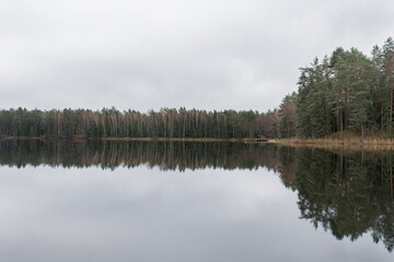 The calm lake reflects the lush green trees and fluffy clouds above, creating a tranquil atmosphere in a forested area during autumn