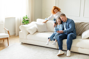 Positive parents and their child girl taking family selfie, using cellphone sitting on comfortable...