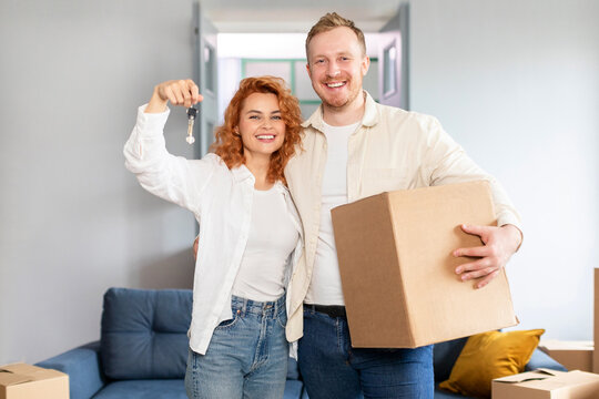 House ownership concept. Happy spouses moving in new apartment, woman showing keys, man holding cardboard box. Insurance, real estate, mortgage concept