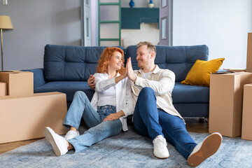 Happy wife and husband sitting on floor near cardboard boxes, giving high five congratulating each...