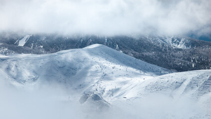 Obraz premium Snowy forest landscape and mountains emerging through holes in a thick layer of cloud