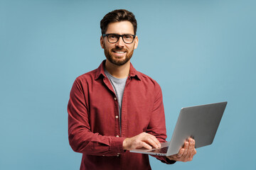 Smiling, happy man wearing eyeglasses, holding laptop, using computer, isolated on blue background