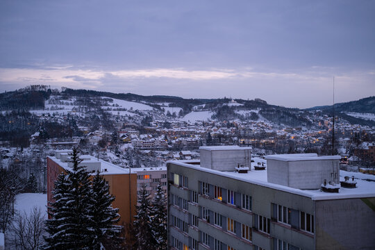 A serene winter view of a European town at dusk. Snow-covered apartment buildings with warm glowing windows, hills, and trees create a cozy atmosphere under an overcast sky.