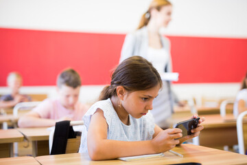 Concentrated diligent tween girl sitting at lesson in classroom, using mobile phone ..