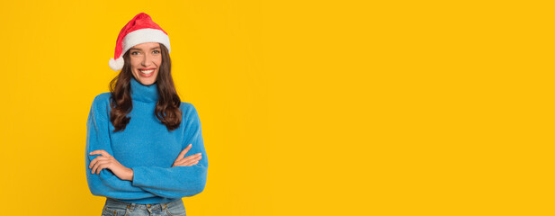 A cheerful woman wearing a Santa hat and a blue sweater stands with her arms crossed, smiling brightly against a vibrant yellow background, embodying festive spirit and joy, copy space