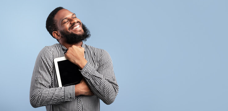 Portrait of emotional black guy hugging tablet with blank screen, holding it tight near chest, blue studio wall