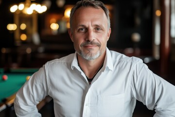 Fototapeta premium Middle-aged man in a white shirt, with a confident expression, posing in an elegant room with blurred background lighting and a pool table insight.