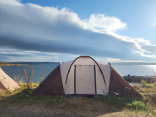 Tents are pitched near a serene lake under expansive skies filled with clouds at sunset. The...
