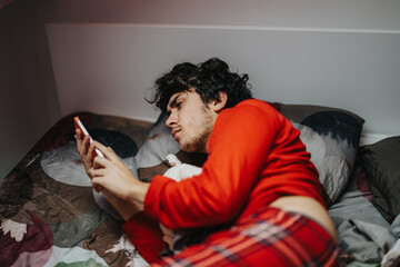 A young man in red pajamas lies in bed, focused on his smart phone. The setting suggests relaxation, comfort, and modern connectivity.