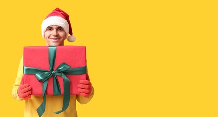 Handsome young man in Santa hat holding Christmas gift on yellow background with space for text