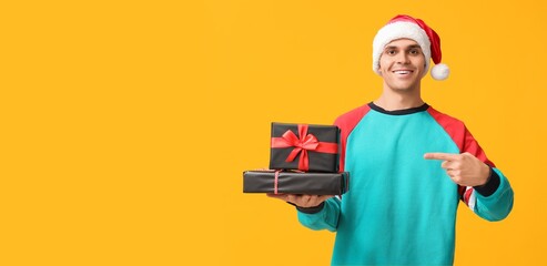Handsome young man in Santa hat holding Christmas gifts on yellow background with space for text