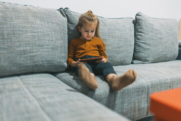 The image captures a curious child captivated by her smartphone while lounging on a couch, set in a...