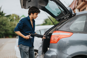 A young man dressed casually places an umbrella and keys into the trunk of a car. The overcast weather and open trunk suggest preparation for a rainy day.