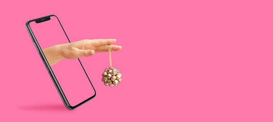 Hand with Christmas decoration coming out of phone screen on pink background with space for text