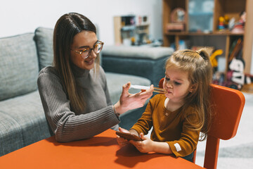 A mother feeds her young daughter while the child is focused on a smartphone at a bright orange table, capturing the blend of technology and parenting.