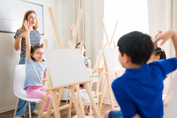 Group of children raising hands and participating in painting lesson with their art teacher at...