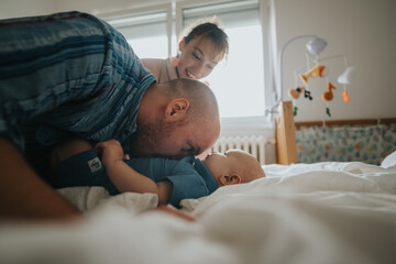 A joyful family moment as parents smile and interact with their baby on a bed. The warm and cozy...