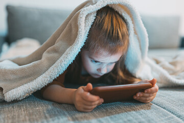 A young child lies engrossed under a fluffy blanket, engaging with a tablet on a couch, reflecting...
