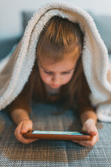 A little girl draped with a fluffy blanket is closely watching her tablet, signifying the influence...