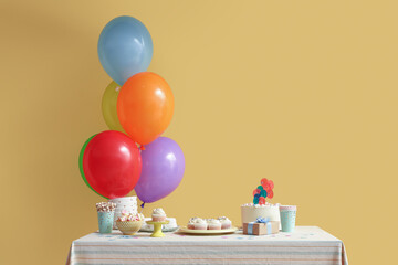 Table with different sweets, gift and balloons for birthday party against yellow wall